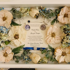 May include: A white frame with a wedding invitation inside. The invitation is for Cecilia Ponce and Gonzalo Campos. The invitation is surrounded by dried flowers, including white roses, blue hydrangeas, and green foliage.