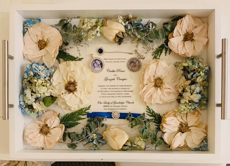 May include: A white frame with a wedding invitation inside. The invitation is for Cecilia Ponce and Gonzalo Campos. The invitation is surrounded by dried flowers, including white roses, blue hydrangeas, and green foliage.