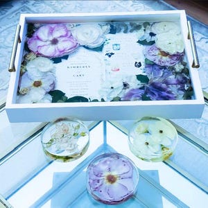 May include: A white tray with gold handles, filled with pressed flowers and a wedding invitation card that reads "Kimberly & Gary". Three smaller trays with pressed flowers are on a glass surface.
