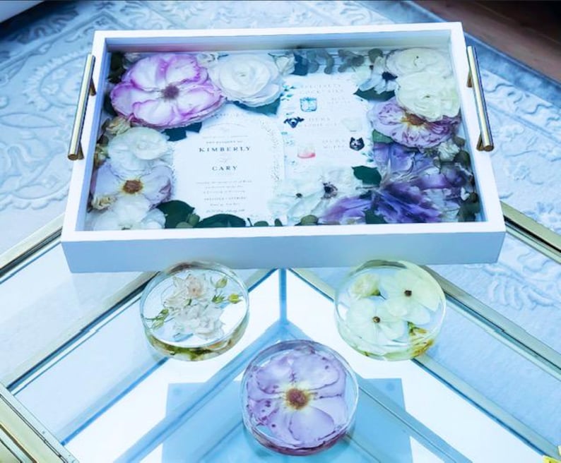 May include: A white tray with gold handles, filled with pressed flowers and a wedding invitation card that reads "Kimberly & Gary". Three smaller trays with pressed flowers are on a glass surface.