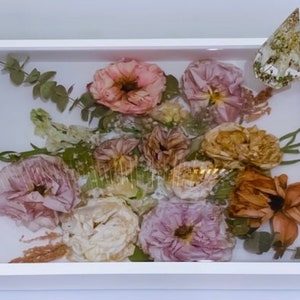 May include: A white tray with gold handles, filled with a resin-sealed arrangement of dried flowers and greenery. The flowers are in shades of pink, peach, and brown, and the greenery is a light green.
