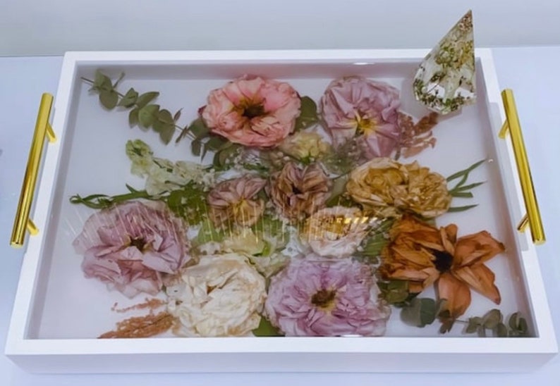 May include: A white tray with gold handles, filled with a resin-sealed arrangement of dried flowers and greenery. The flowers are in shades of pink, peach, and brown, and the greenery is a light green.