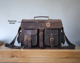 Op de afbeelding: Een bruine leren schoudertas met een voorflap en meerdere vakken. De tas heeft een messing gesp en een band met een messing sluiting. De tas is gepersonaliseerd met de naam "JAMES" gegraveerd op de voorkant.
