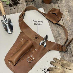 May include: A brown leather gardening tool belt featuring multiple pockets, a metal D-ring, and an adjustable strap. The leather is embossed with a circular logo above the text Engraving Sample, and a pair of metal pruning shears is tucked inside.