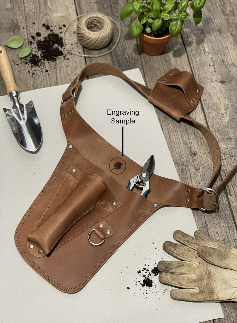May include: A brown leather gardening tool belt featuring multiple pockets, a metal D-ring, and an adjustable strap. The leather is embossed with a circular logo above the text Engraving Sample, and a pair of metal pruning shears is tucked inside.