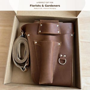 May include: A brown leather tool holster with a matching adjustable strap, featuring multiple pockets, silver-toned metal rivets, and a D-ring. The item is presented inside a cardboard box with the text A PERFECT GIFT FOR Florists & Gardeners.