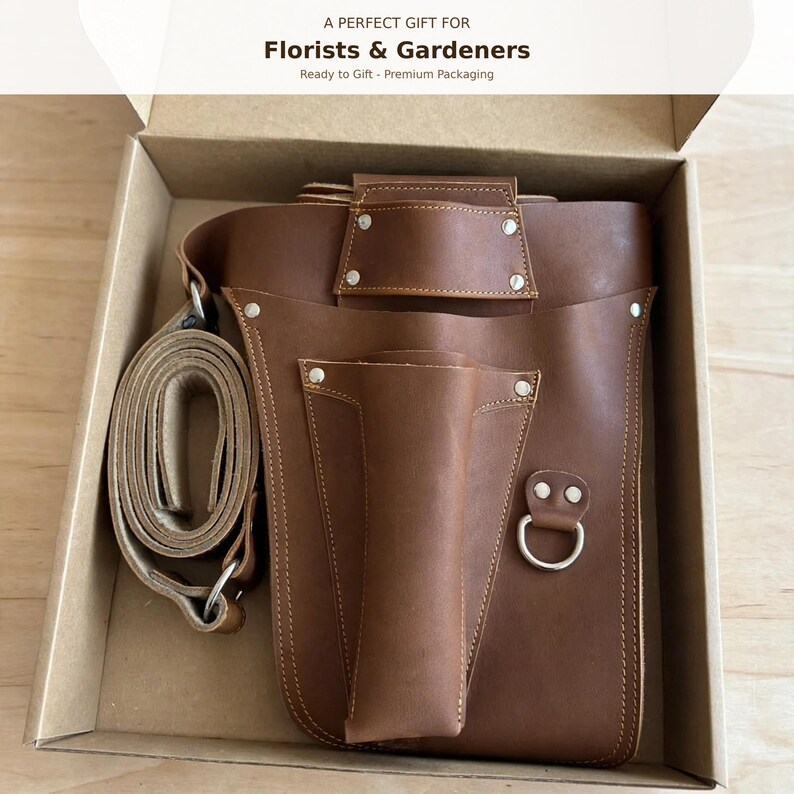 May include: A brown leather tool holster with a matching adjustable strap, featuring multiple pockets, silver-toned metal rivets, and a D-ring. The item is presented inside a cardboard box with the text A PERFECT GIFT FOR Florists & Gardeners.