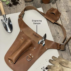 May include: A brown leather gardening tool belt featuring multiple pockets, a metal D-ring, and an adjustable strap. The leather is embossed with the text "Engraving Sample" and the name "ALBERT" above a pair of metal pruning shears.