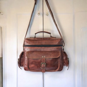May include: A brown leather camera bag with a long adjustable strap, a front flap with a buckle closure, and two side pockets.