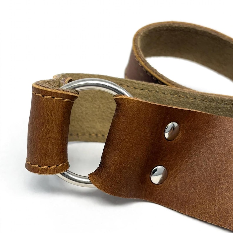 May include: A brown leather strap featuring a smooth, circular silver-toned metal ring attachment. The leather is secured to the ring with visible stitching and two polished silver-toned metal rivets, showcasing the natural texture of the material.