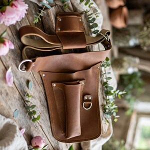 May include: A brown leather tool holster featuring a belt loop attachment, a vertical pocket for tools, and a metal D-ring. The holster is constructed with visible stitching and silver-toned metal rivets securing the leather panels together.