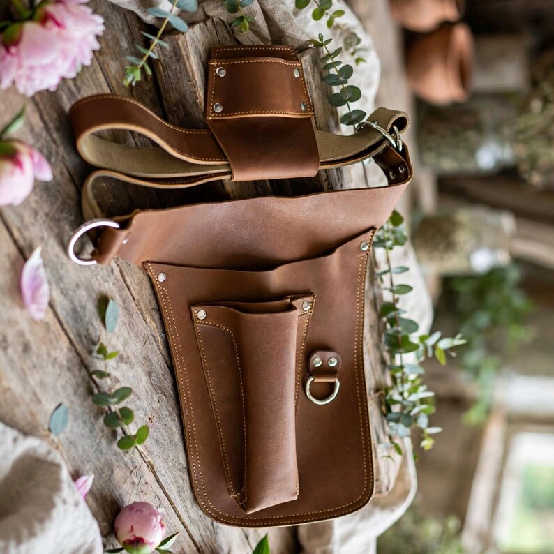 May include: A brown leather tool holster featuring a belt loop attachment, a vertical pocket for tools, and a metal D-ring. The holster is constructed with visible stitching and silver-toned metal rivets securing the leather panels together.