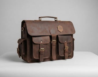 May include: A dark brown leather messenger bag featuring a top handle, a large front flap with a central buckle strap, two exterior front pockets with buckle closures, side pockets, and a small leather tag attached to the handle with a string.
