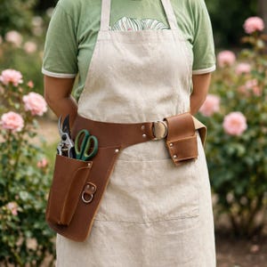 May include: A beige linen apron worn over a green t-shirt, featuring a brown leather tool belt with a large holster containing green-handled shears and a smaller leather pouch attached to the side with a metal ring and silver rivets.