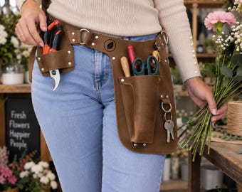 May include: A brown leather tool belt worn around the waist, featuring multiple pockets and loops. The belt includes a small leaf logo, metal rivets, and a metal ring for keys. It holds various gardening tools, including orange and black pruners.