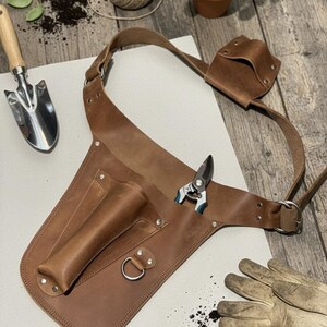 May include: A brown leather gardening tool belt featuring multiple pockets, a metal D-ring, and an adjustable strap. The belt is designed to hold various gardening implements, with a pair of pruning shears tucked into one of the main storage slots.