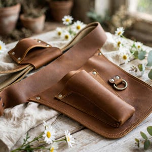 May include: A brown leather tool belt featuring multiple pockets, reinforced silver-tone metal rivets, and a circular metal ring attachment. The belt is constructed from smooth, matte-finish leather with visible stitching along the edges and pockets.
