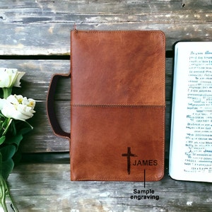 May include: A brown leather Bible cover with a cross and the name "JAMES" engraved on the front. The cover has a handle and a stitched seam.
