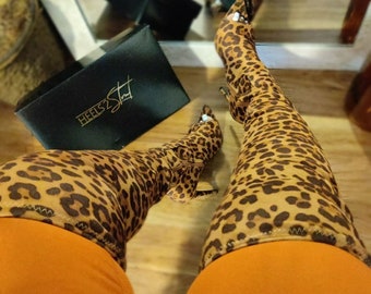 Puede incluir: Un par de botas altas de leopardo marrón y negro con punta puntiaguda y tacón de aguja. Las botas se llevan con leggings naranjas y el texto "HEELS2STUN" es visible en una caja negra.