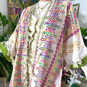 May include: A beige kimono-style jacket with a colorful geometric pattern in pink, blue, yellow, and green. The jacket has white sleeves with green trim and tassels. The outfit includes beige pants and a white top.