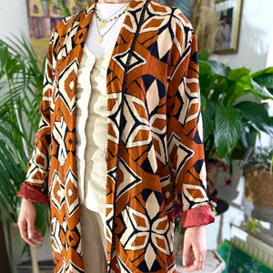 Op de afbeelding: Een lange, open jas met patroon in bruin, crème, zwart en marineblauw. De jas heeft een geometrisch ontwerp en rode manchetten. Het kledingstuk wordt gedragen over een crèmekleurige outfit.