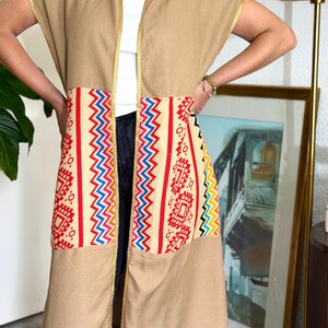 Op de afbeelding: Een mouwloos beige lang vest met een crèmekleurig middengedeelte met rode, blauwe en gele geometrische patronen. Het kledingstuk heeft gouden biezen langs de opening aan de voorzijde en de kraag, en een donkerblauwe franje aan de onderzoom.