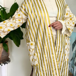 May include: A long-sleeved open-front jacket featuring a vertical gold and white striped pattern on the main body. The sleeves and two front patch pockets display a contrasting white fabric with a repeating yellow and gold fan-shaped floral motif.
