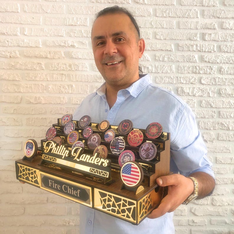 Op de afbeelding: Een houten displaystandaard met een gouden naamplaatje waarop "Phillip Landers Fire Chief" staat. De standaard heeft meerdere niveaus om ronde, kleurrijke challenge-munten te tonen. De standaard wordt door een persoon vastgehouden.