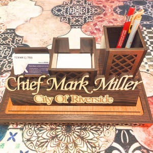 May include: A wooden desk organizer with a nameplate that reads "Chief Mark Miller City of Riverside". The organizer has a pen holder, a business card holder, and a note pad holder.