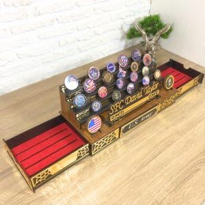 Op de afbeelding: Een houten displaykast met een lade bekleed met rood vilt. De kast is ontworpen om een ​​collectie challenge-munten te bewaren en te presenteren. De kast heeft een naamplaatje met de tekst "SFC David Taylor" en een plaquette met de tekst "U.S. Army".