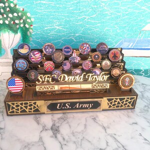 Op de afbeelding: Een houten displaykast met een gouden rand en een zwarte achtergrond. De kast is gevuld met een verzameling challenge-munten. De kast heeft een naamplaatje met de tekst "SFC David Taylor" en een plaquette met de tekst "U.S. Army".