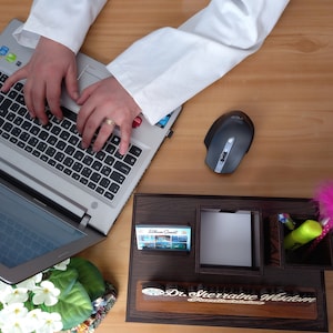 May include: A wooden desk organizer with compartments for pens, pencils, and a notepad. The organizer has a nameplate that reads "Dr. Sherraine Wisdom". A wireless mouse, a pink feather pen, and a laptop are also on the desk.