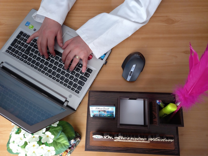 May include: A wooden desk organizer with compartments for pens, pencils, and a notepad. The organizer has a nameplate that reads "Dr. Sherraine Wisdom". A wireless mouse, a pink feather pen, and a laptop are also on the desk.