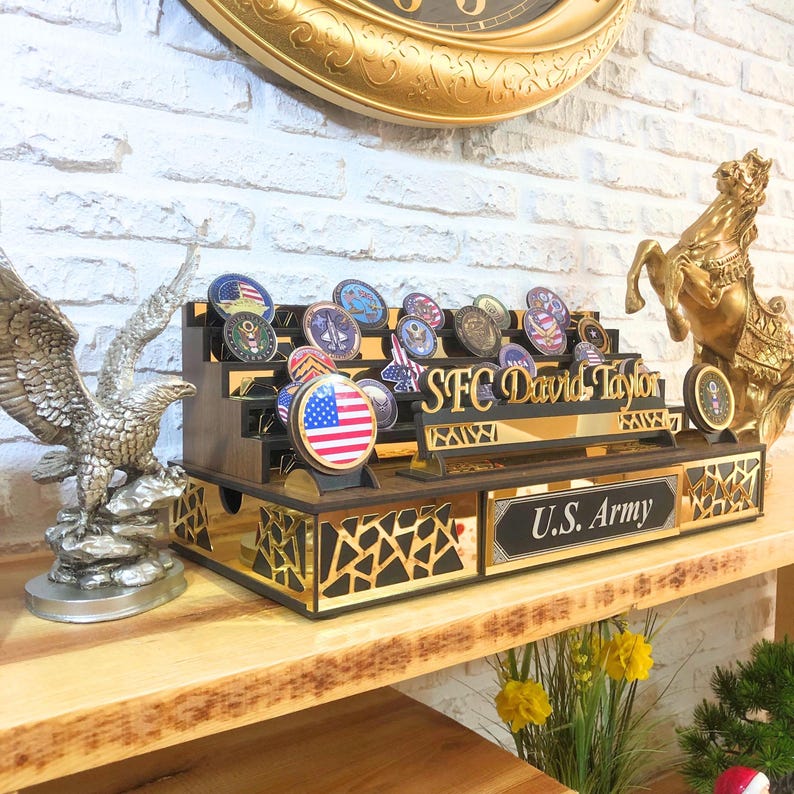 Op de afbeelding: Een houten displaystandaard met een gouden afwerking, met een gepersonaliseerd naamplaatje met de tekst "SFC David Taylor" en een kleiner plaatje met de tekst "U.S. Army". De standaard is versierd met verschillende militaire patches en een kleine Amerikaanse vlag.