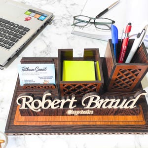 May include: A wooden desk organizer with a nameplate that reads "Robert Brand" and the word "Captain" underneath. The organizer has a compartment for pens, a compartment for sticky notes, and a business card holder. The business card holder has a business card that reads "Tutkum Sanat".