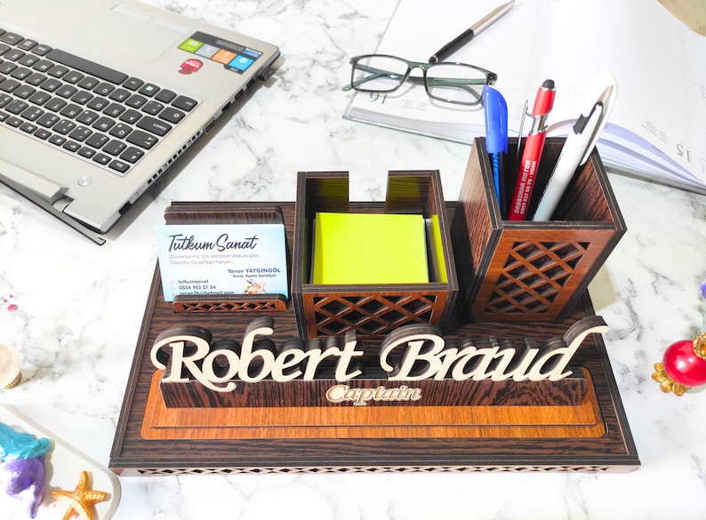 May include: A wooden desk organizer with a nameplate that reads "Robert Brand" and the word "Captain" underneath. The organizer has a compartment for pens, a compartment for sticky notes, and a business card holder. The business card holder has a business card that reads "Tutkum Sanat".