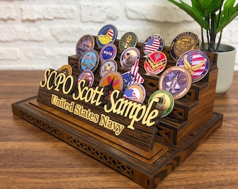 May include: A tiered wooden display stand for challenge coins featuring the text SCPO Scott Sample United States Navy in gold lettering. The stand holds numerous circular military and space-themed coins with various colorful emblems and insignias.