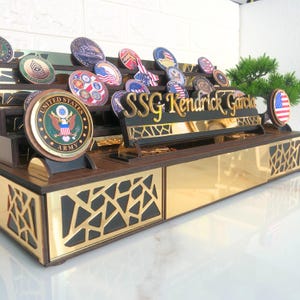 Op de afbeelding: Een houten displaystandaard met een gouden afwerking, met een verzameling militaire en NASA-challenge-munten. De standaard bevat ook een naamplaatje met de tekst "SSG Kendrick Garcia".