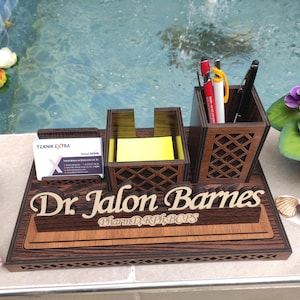 May include: A wooden desk organizer with a nameplate that reads "Dr. Jalon Barnes, PharmD, RPh, BCPS". The organizer has a pen holder, a note pad holder, and a business card holder. The organizer is made of dark wood with a wood grain pattern.