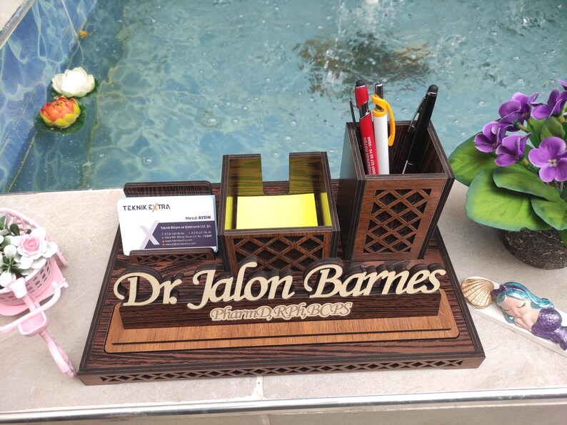 May include: A wooden desk organizer with a nameplate that reads "Dr. Jalon Barnes, PharmD, RPh, BCPS". The organizer has a pen holder, a note pad holder, and a business card holder. The organizer is made of dark wood with a wood grain pattern.