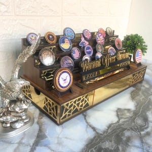 Op de afbeelding: Een houten displaykast met een gouden rand en een zwart adelaarbeeldje. De kast heeft meerdere planken om ronde, kleurrijke militaire challenge-munten te tonen. Een houten bordje met de tekst "Warrant Officer Brown".