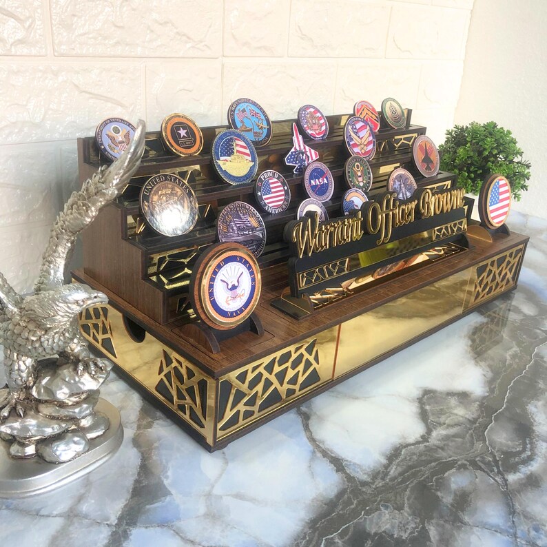 Op de afbeelding: Een houten displaykast met een gouden rand en een zwart adelaarbeeldje. De kast heeft meerdere planken om ronde, kleurrijke militaire challenge-munten te tonen. Een houten bordje met de tekst "Warrant Officer Brown".