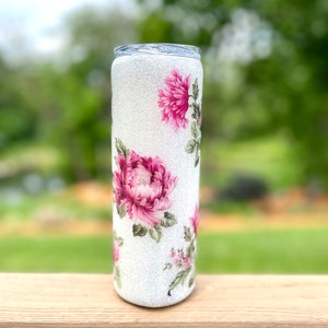 May include: A white glitter tumbler decorated with pink peony floral designs and green leaves. The tumbler has a clear lid and is cylindrical. The floral pattern is repeated around the tumbler.