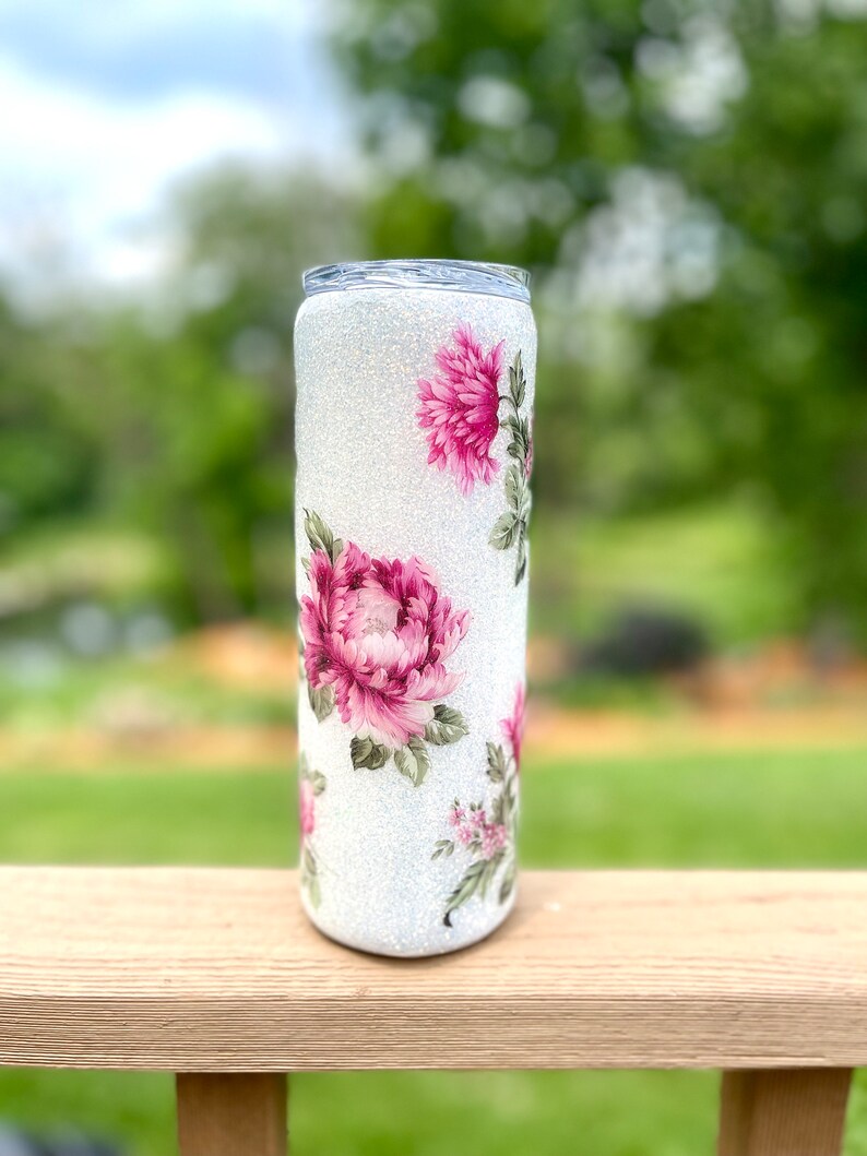 May include: A white glitter tumbler decorated with pink peony floral designs and green leaves. The tumbler has a clear lid and is cylindrical. The floral pattern is repeated around the tumbler.