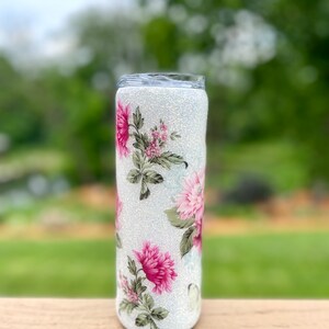 May include: A glitter-covered tumbler with a floral design. The tumbler has a white, sparkling base and is decorated with pink and green floral patterns. The design wraps around the entire tumbler, creating a decorative and eye-catching look.