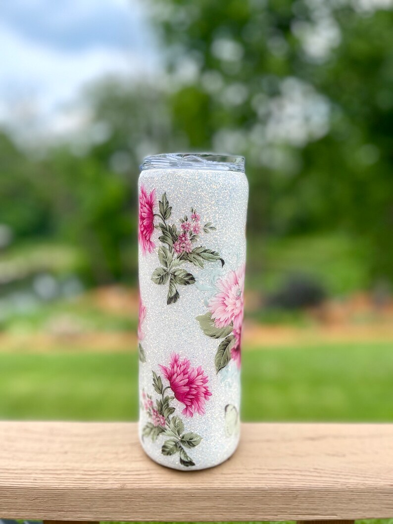 May include: A glitter-covered tumbler with a floral design. The tumbler has a white, sparkling base and is decorated with pink and green floral patterns. The design wraps around the entire tumbler, creating a decorative and eye-catching look.