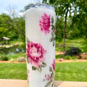May include: A white glitter tumbler with pink floral patterns. The tumbler has a cylindrical shape and a silver lid. The floral design includes large pink peonies and smaller pink flowers with green leaves. The background is a blurred outdoor scene.
