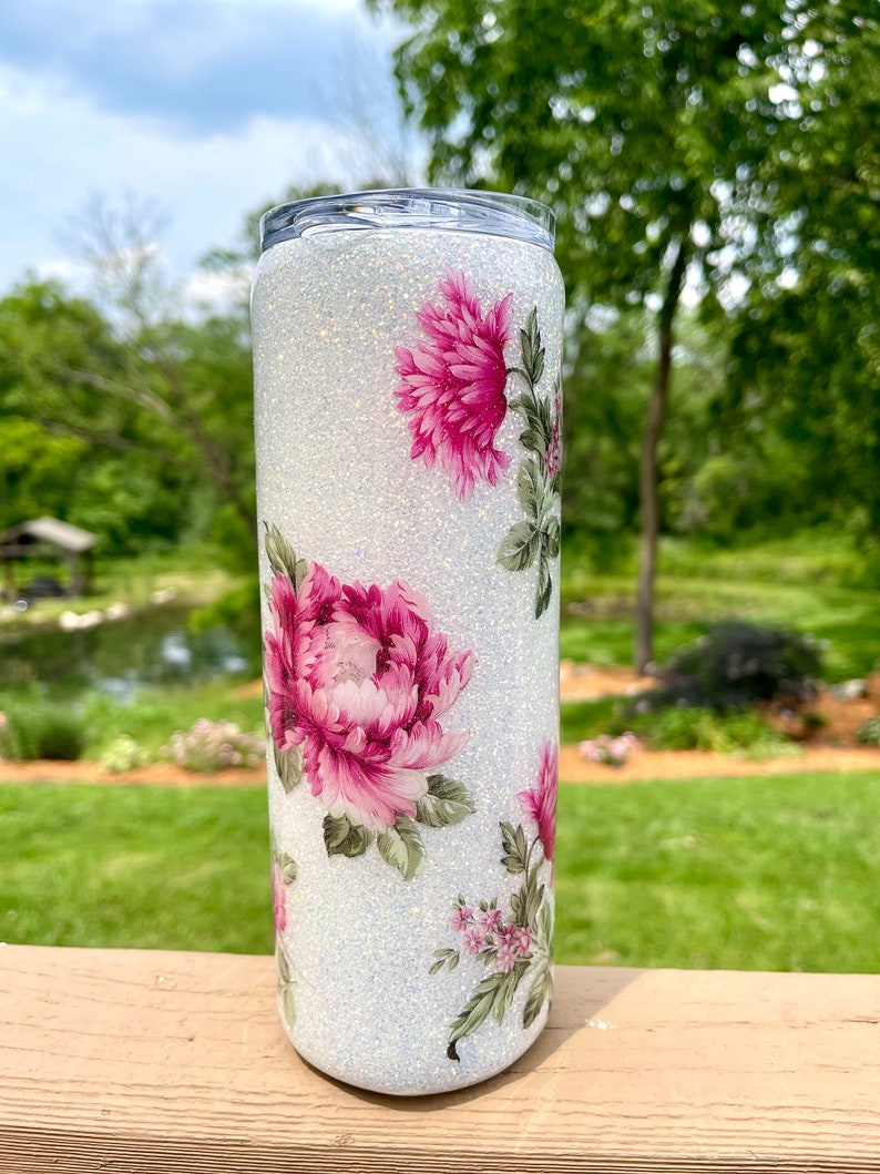 May include: A white glitter tumbler with pink floral patterns. The tumbler has a cylindrical shape and a silver lid. The floral design includes large pink peonies and smaller pink flowers with green leaves. The background is a blurred outdoor scene.