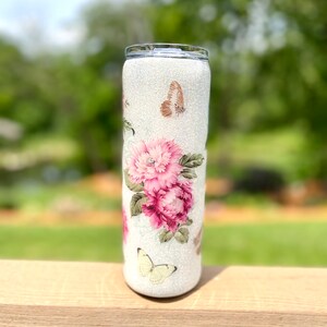 May include: A white glitter tumbler decorated with pink peonies and butterflies. The floral design features a cluster of large, vibrant pink flowers with green leaves. Delicate butterflies in shades of brown and yellow are scattered around the tumbler.