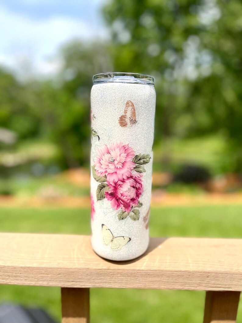 May include: A white glitter tumbler decorated with pink peonies and butterflies. The floral design features a cluster of large, vibrant pink flowers with green leaves. Delicate butterflies in shades of brown and yellow are scattered around the tumbler.